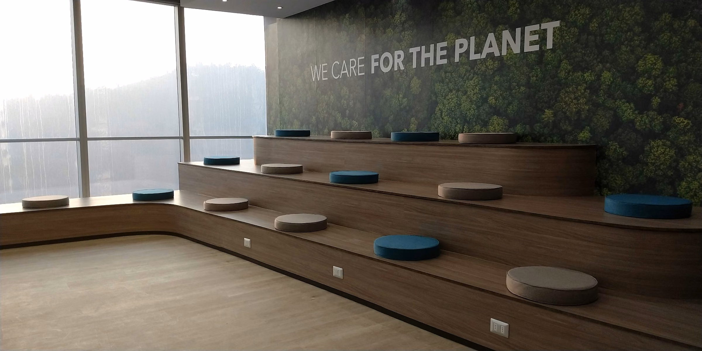 Auditorio interior con gradas de madera y cojines tapizados frente a un muro verde, Oficina Titanium